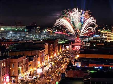 Nighttime cityscape of Nashville with a large fireworks display over buildings and a crowd.