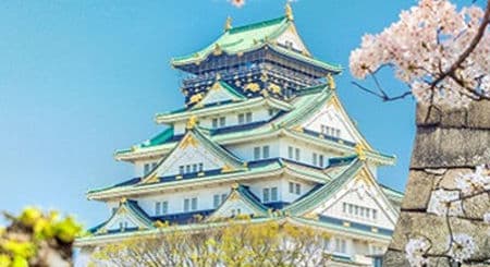 Grand Japanese castle with green roofs under blue sky, surrounded by delicate cherry blossoms.