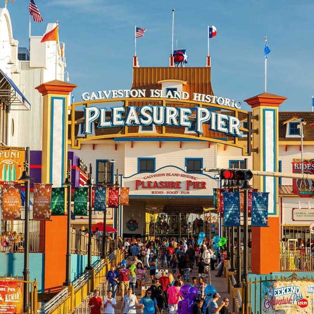 Galveston Island Historic Pleasure Pier entrance archway with crowds and flags under blue sky.